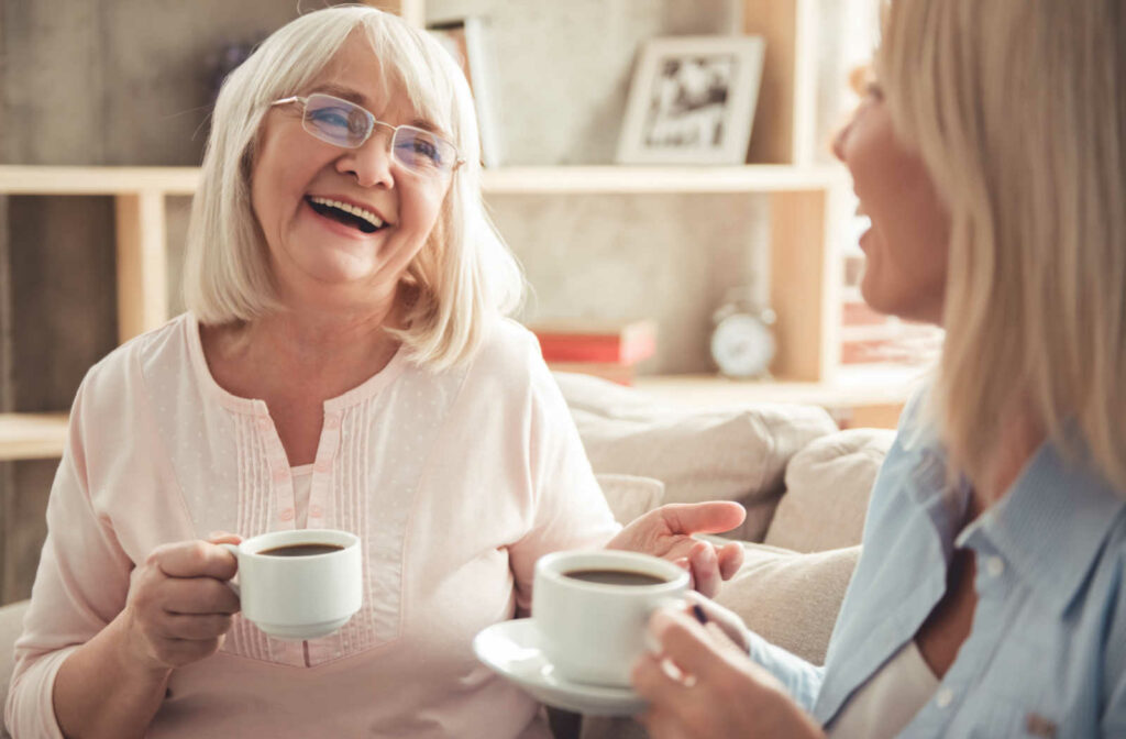 A senior woman and her daughter sitting on a couch smiling and talking to each other while holding a cup of tea.