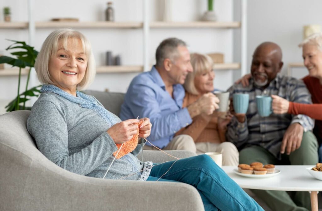 An older adult smiles while knitting an orange scarf, while their friends in the background clink coffee mugs together